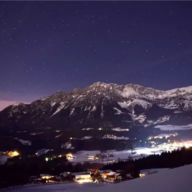 Eine winterliche Landschaft bei Nacht mit schneebedeckten Bergen und einem klaren Sternenhimmel. Kleine Lichter von Dörfern leuchten sanft im Schnee.