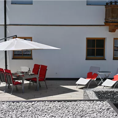 Ein gemütlicher Außenbereich mit einem Tisch und roten Stühlen unter einem Sonnenschirm. Es gibt auch zwei Liegen und eine moderne Umgebung aus Steinen.