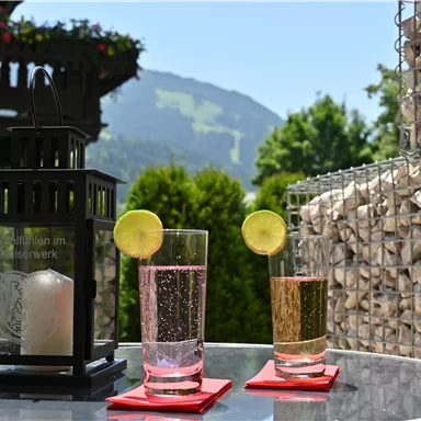 Ein Tisch im Freien mit zwei Gläsern Getränken und einer Laterne. Im Hintergrund sind grüne Berge und ein klarer Himmel zu sehen.