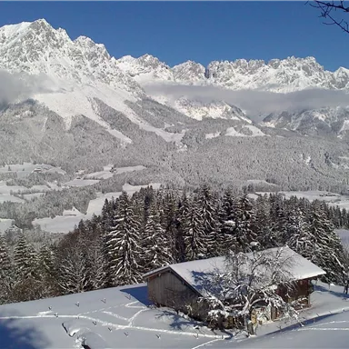 A snow-covered landscape with high mountains in the background. In the foreground is a small cabin, surrounded by trees and snow.