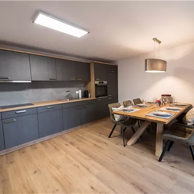 A modern kitchen with a large dining table and elegant chairs. The walls are in neutral colors, and the floor is made of wood.