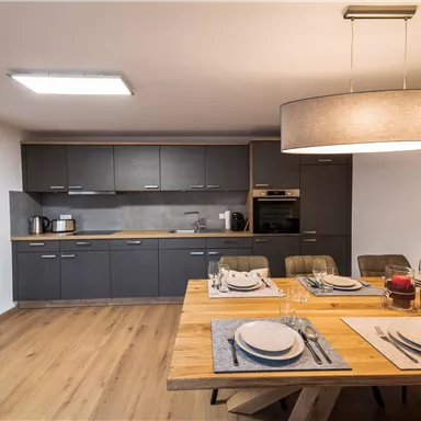 A modern kitchen with a large wooden dining table. The walls are in a neutral color tone, and the furnishings are elegant and functional.