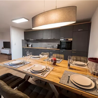 A modern dining room with a large wooden table and stylish chairs. The open kitchen in the background is functional and well-equipped.