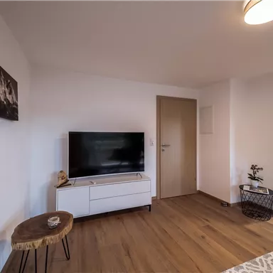 A modern living room with a TV and a wooden table. The walls are light, and the floor is made of wood.