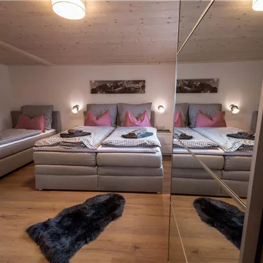A modern bedroom with multiple beds and cozy furnishings. There are bright walls, soft lighting, and a warm wooden floor.