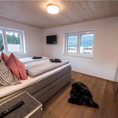 A bright bedroom with a large bed and several pillows. There are two windows, a television on the wall, and a soft carpet on the floor.