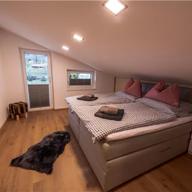 A cozy bedroom with a double bed and two pillows. Large windows provide daylight, and the floor is wooden.