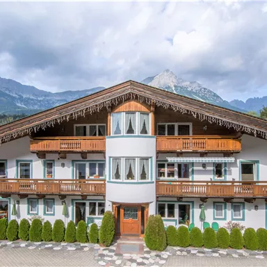 Ein traditionelles Alpenhaus mit dekorativen Balkonen und viel Grün umgeben. Im Hintergrund sind majestätische Berge und ein bewölkter Himmel zu sehen.