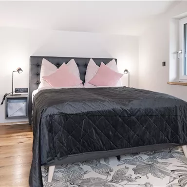 A modern bedroom with a black bed and pink pillows. The walls are light and there is a window with natural light.