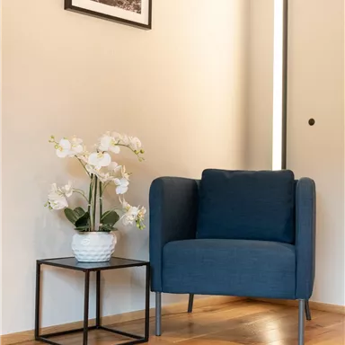 A cozy blue armchair stands next to a small table with a white orchid. A picture hangs on the wall, and there is a warm glow in the background.