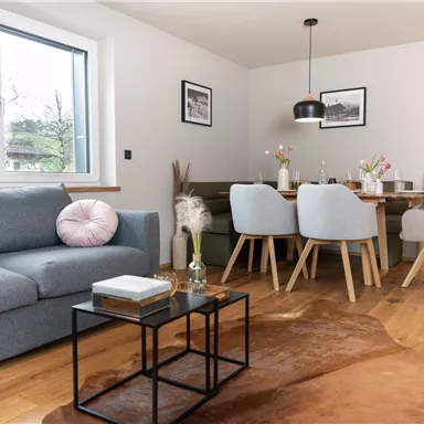 A modern living room with a gray sofa and a small table. In the background, there is a dining table with chairs and decorative elements.