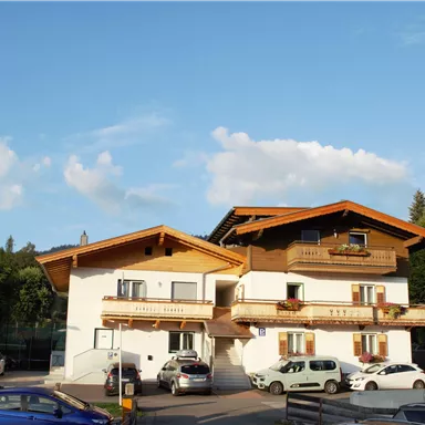 A comfortable building in alpine style with wooden cladding and balconies. In front of the house, several cars are parked under a clear blue sky.