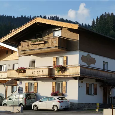 A traditional alpine house with two floors and a wooden porch. Several cars are parked in front of the house.