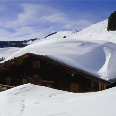 A cozy wooden house is surrounded by deep snow. In the background, snow-covered mountains can be seen under a clear blue sky.