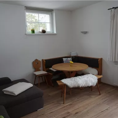 Ein gemütlicher Wohnraum mit einem runden Esstisch und einer Sitzgelegenheit. Der Holzfußboden und das natürliche Licht sorgen für eine einladende Atmosphäre.