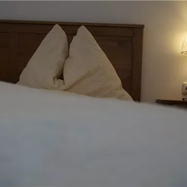 Ein gemütliches Schlafzimmer mit einem Holzbett und Kissen. Eine sanfte Lampe neben dem Bett sorgt für eine entspannte Atmosphäre.