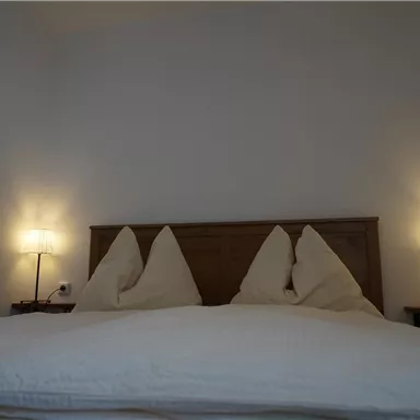 Ein gemütliches Schlafzimmer mit einem großen Bett und zwei Nachttischen. Die Kissen sind in einer schönen Form, und sanftes Licht von den Lampen sorgt für eine angenehme Atmosphäre.