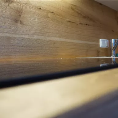 Eine moderne Küchenarbeitsplatte aus Holz mit einem glänzenden Bereich. Im Hintergrund sind ein Wasserhahn und eine Steckdose sichtbar.