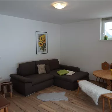 Ein gemütliches Wohnzimmer mit einem braunen Sofa und einem runden Tisch. An der Wand hängt ein Bild von Sonnenblumen, und ein Fenster lässt viel Tageslicht herein.