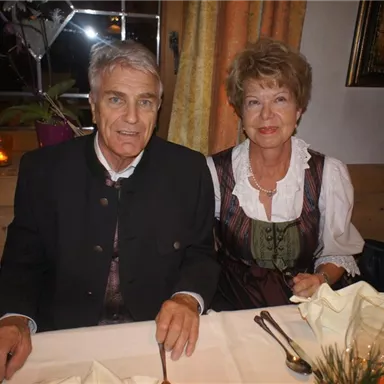Ein Paar in traditioneller Tracht sitzt an einem gedeckten Tisch. Im Hintergrund sind dekorative Vorhänge und eine warm beleuchtete Atmosphäre zu sehen.
