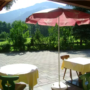 A cozy outdoor area with two tables and a sunshade. The view of the green landscape and the mountains in the background creates a relaxed atmosphere.
