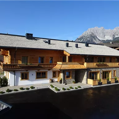 A beautiful wooden house in the Alps with a steep roof. In the background, impressive mountains and a clear blue sky can be seen.