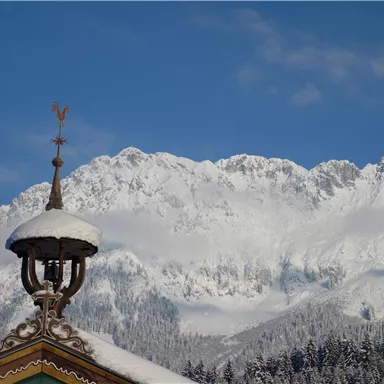 Eine verschneite Berglandschaft mit hohen Gipfeln und klarem Himmel. Im Vordergrund ist ein traditionelles Dach mit einer Wetterfahne zu sehen.