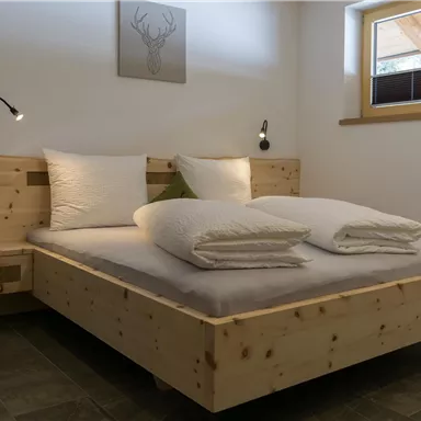 A cozy bedroom with a wooden bed and two large, white pillows. There is soft lighting and a window with a view of the outside world.