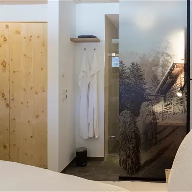 A cozy bedroom with a wooden wardrobe and a modern bathroom. The walls are light, while the glass door features a beautiful mountain motif.