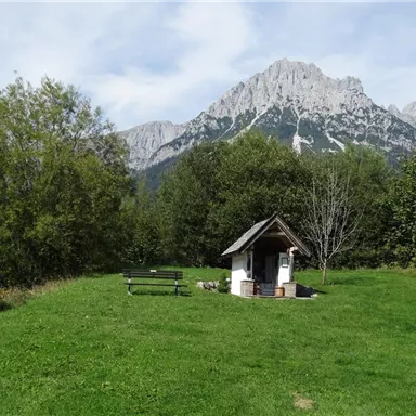 Eine malerische Landschaft mit einem kleinen Häuschen und einer Bank auf einer Wiese. Im Hintergrund ragen majestätische Berge in den Himmel.