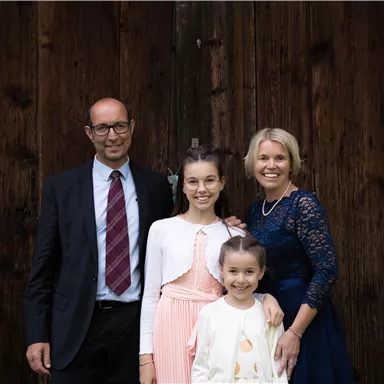Eine Familie steht vor einer Holzwand. Sie lächeln freundlich und tragen festliche Kleidung.
