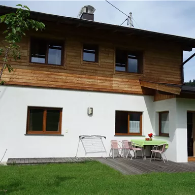 Ein hübsches Haus mit einem Holzverkleidung und weißer Fassade. Im Vordergrund gibt es eine Terrasse mit einem Tisch und Stühlen.