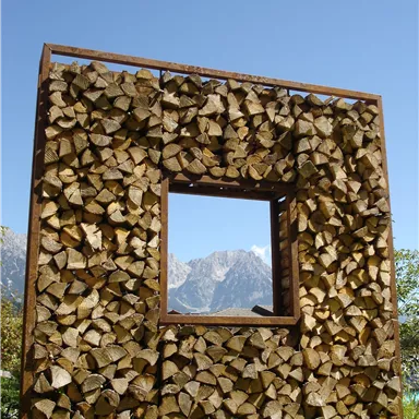 A wooden frame with a rectangular cutout, filled with firewood. In the background, there are mountains and a clear blue sky.