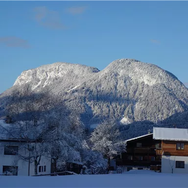 Eine Winterlandschaft mit schneebedeckten Bergen und klar blauem Himmel. Im Vordergrund sind einige Häuser umgeben von Schnee zu sehen.