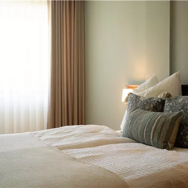 Ein gemütliches Schlafzimmer mit einem einladenden Bett und weichen Kissen. Das sanfte Licht der Fenster schafft eine entspannte Atmosphäre.