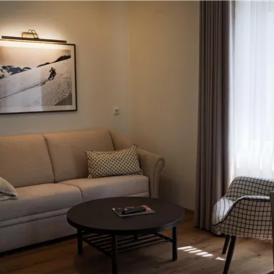 Ein gemütliches Wohnzimmer mit einem hellen Sofa und einem modernen Sessel. Ein runder Tisch und ein großes Fenster sorgen für viel Tageslicht.