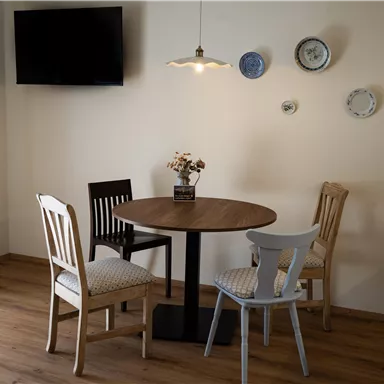 Ein gemütlicher Esstisch aus Holz mit drei Stühlen in einem hellen Raum. An der Wand hängen einige Teller und ein Fernseher ist sichtbar.