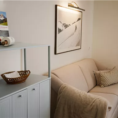 Ein gemütliches Wohnzimmer mit einem beigen Sofa und einem kleinen Tisch. An der Wand hängt ein Schwarz-Weiß-Foto von einer Berglandschaft.