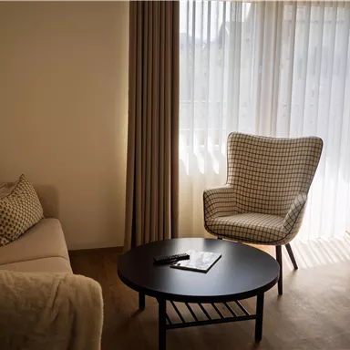 Ein gemütlicher Wohnbereich mit einem hellen Sofa und einem bequemen Sessel. Das Licht strömt durch die Vorhänge und schafft eine einladende Atmosphäre.