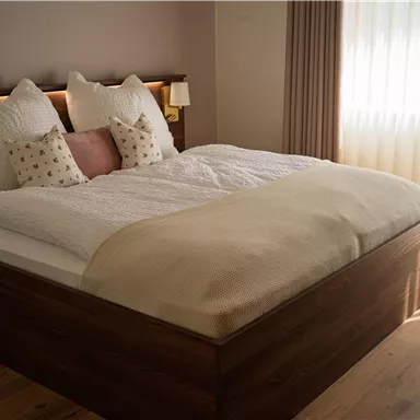 Ein modernes Schlafzimmer mit einem großen Bett und flauschigen Kissen. Weiches Licht fällt durch die Fenster und sorgt für eine gemütliche Atmosphäre.