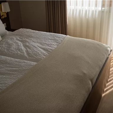 Ein gemütliches Schlafzimmer mit einem großen Bett und weichen Decken. Tageslicht strömt durch die Vorhänge und wirft interessante Schatten auf den Boden.