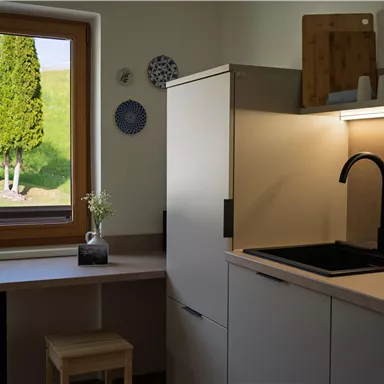 Eine moderne Küche mit einer hellen Arbeitsfläche und einem schwarzen Waschbecken. Durch das Fenster sind grüne Bäume und eine sonnige Landschaft zu sehen.