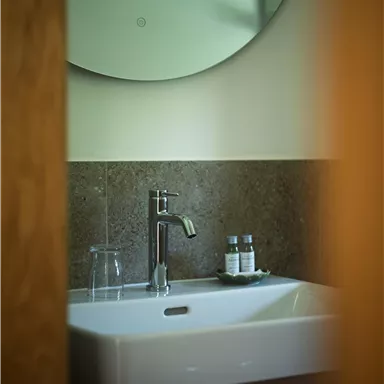 Ein elegantes Badezimmer mit einem weißen Waschbecken und einem modernen Wasserhahn. Ein runder Spiegel und ein Glas Wasser ergänzen das stilvolle Ambiente.