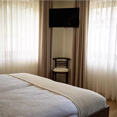Ein helles Schlafzimmer mit einem großen Bett und einem Fernseher an der Wand. Neben dem Bett steht ein einzelner Stuhl und große, helle Vorhänge lassen das Licht herein.