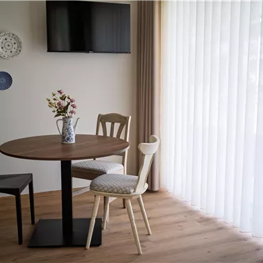 Ein gemütlicher Essbereich mit einem runden Tisch und drei Stühlen. An der Wand hängen dekorative Teller, und große Fenster lassen viel Licht herein.