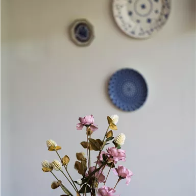 Eine elegante Kanne mit bunten Blumen steht auf einem Tisch. Im Hintergrund hängen dekorative Teller an der Wand.