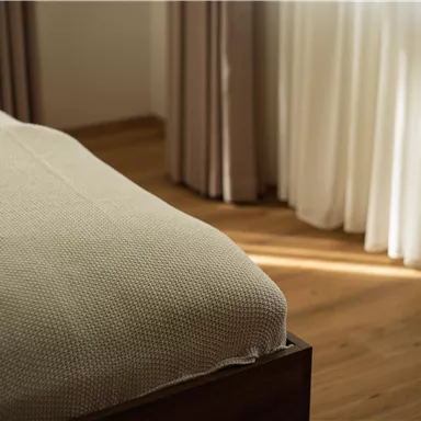 Ein gemütliches Schlafzimmer mit einem Bett und einer hellen, neutralen Decke. Weiche Vorhänge lassen warmes Licht herein.