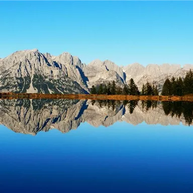 Eine malerische Berglandschaft mit klaren Gewässern, die die Gipfel widerspiegeln. Der Himmel ist strahlend blau und vermittelt eine ruhige Atmosphäre.