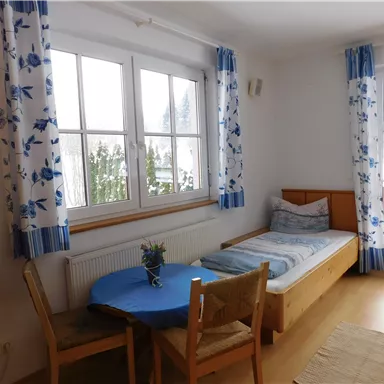 Ein helles Zimmer mit einem Einzelbett und einem Tisch. Große Fenster lassen viel Licht herein.