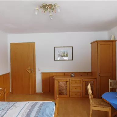 Ein gemütliches Zimmer mit Holzmöbeln und einem kleinen Tisch. An der Wand hängt ein Bild, und die Fenster sind mit blauen Vorhängen dekoriert.